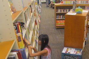 A young patron reviews the selection of books available in the children's section of the Washougal Community Library. There are discussions regarding the relocation of the library to a larger, existing building or a new structure.