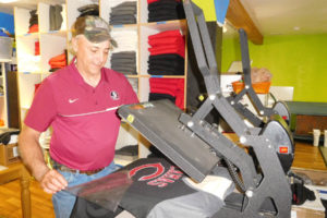 Dave Gray, co-owner of Glory Days Trophies & Sports, uses a heat press to apply a Camas High School emblem to a T-shirt. Glory Days also sells clothing and accessories that promote Washougal High School. 