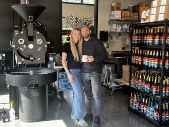 Camas natives Anita and Ben Mikhalets stand between Ben&rsquo;s coffee roaster and a shelf of espresso drink flavors inside the couple&rsquo;s newly opened Daily Paper Coffee Roasters roastery and drive-thru cafe, located at 3016 N.E. Everett St., in Camas, on Oct. 10, 2024.